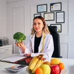 Female nutritionist in office holding broccoli, surrounded by fruits, promoting healthy lifestyle.