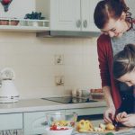 Young pretty mother teaching her cute daughter to cut vegetables properly. Little smiling girl cooking together with loving mom at home in modern kithcen